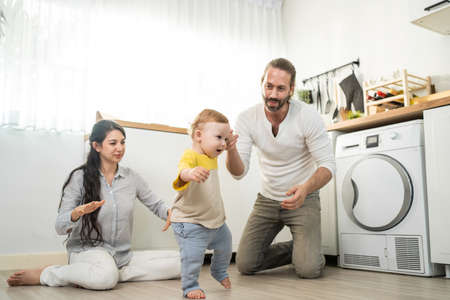 Caucasian baby boy child learn to walk with parents support in house. Happy family, mother and father helping young toddler son taking first step walk on floor to develop skill in living room at home.の写真素材