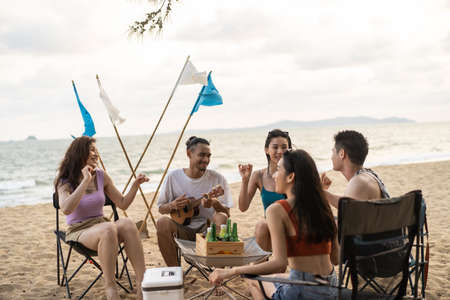 Group of Asian young man and woman having party on the beach together. Attractive happy friends traveler singing and dancing while camping at seaside enjoy holiday vacation trip in tropical sea islandの写真素材