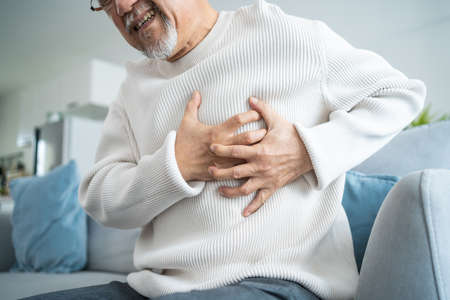 Asian senior older man having chest pain feel suffer from heart attack. Attractive older mature patient has difficulty breath clutching his chest from acute pain while sitting on sofa in living room.の写真素材