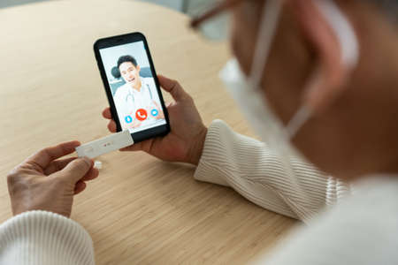 Asian senior man video call with doctor in living room at home. Elderly older man patient consult with specialist practitioner after using ATK rapid test kit to check positive 2 stripe result at home.の写真素材