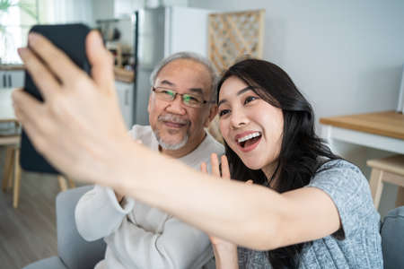 Asian lovely family, young daughter use phone selfie with older father. Beautiful girl and senior elderly male sit on sofa, enjoy video call by internet online talking together in living room at home.の写真素材