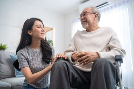 Asian daughter support old disabled man sitting on wheelchair at home. Beautiful girl help and take care of senior elderly mature handicap father patient doing physical therapy in living room in houseの写真素材
