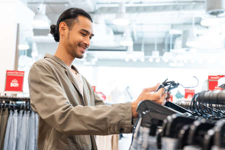 Asian young man look and choose products of clothes in shopping mall. Attractive happy male enjoy walking in department store to choose new t-shirt and pants from clothing racks in marketplace center.の写真素材