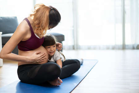 Asian young woman pregnant and little girl doing Yoga Pilates at home. Attractive mother pregnancy with kid daughter in sportswear feel happy and relax, follow exercise instruction from online trainerの写真素材