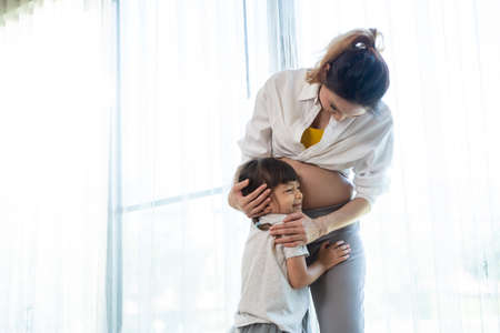 Asian young woman pregnant and little girl standing hugging each other. Attractive mother pregnancy with kid daughter, sister hug baby in womb together with loving and happiness in living room at homeの写真素材