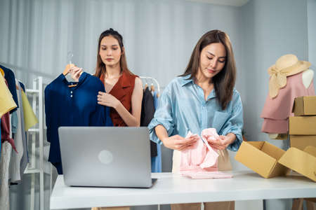 Asian woman friends couple packing clothes order into box for customer. Young attractive business people work to preparing parcel boxes check ecommerce shipping online retail to sell at office store.の写真素材
