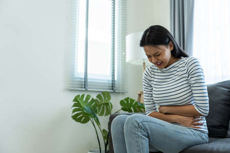 Asian beautiful sick girl suffer from hurt stomachache in living room. Attractive young woman feel bad and painful after wake up on bed, having stomach ache or periods then clasping belly in morning.の写真素材