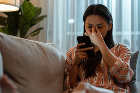 Exhausted Asian young woman hurt eye while using mobile phone at night. Attractive girl sitting on...