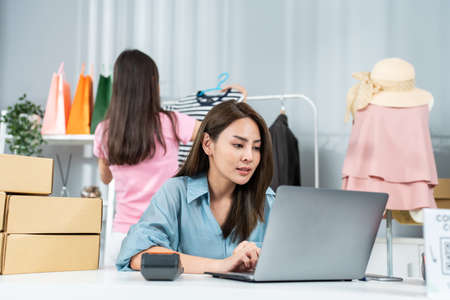 Asian woman friends couple packing clothes order into box for customer. Young attractive business people work to preparing parcel boxes check ecommerce shipping online retail to sell at office store.の写真素材