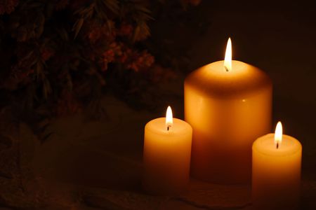 Three burning candles of different sizes in darkness with flowers in the background on a festive...