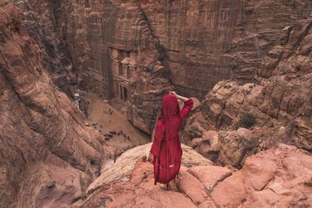 A woman traveller wearing red dress standing on top of mountain looking to the Treasury in Petra...