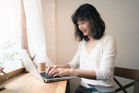 Young Asian woman working and video conferencing with laptop computer. A happy woman with smiley face working from home. A cup of coffee on wooden table. Social distancing concept. Bangkok, Thailandの写真素材