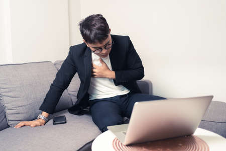 Young Asian business man sitting on sofa and working from home. Man feeling stress and suffering from heart failure. Tired Asian young entrepreneur manager. Work from home concept. Bangkok, Thailandの写真素材