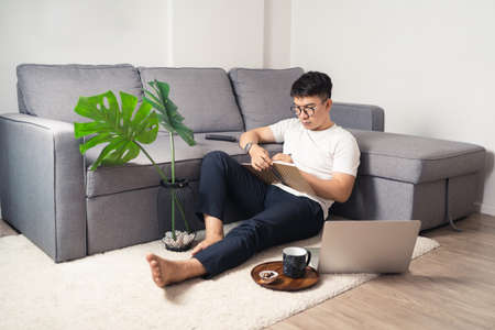 Young Asian man working from home and writing on notebook after working by using laptop computer. Social distancing situation. Work from home concept. Bangkok, Thailandの写真素材