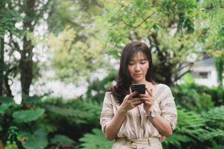 Young Asian woman using smart phone for chating or seaching information. A woman with smiley face in greeny garden. Digital lifestyle concept. Bangkok, Thailandの写真素材