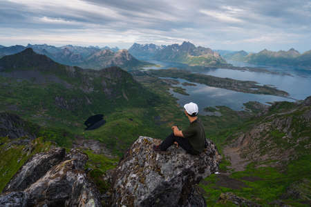Young Asian man trekker sitting on rock on top of Floya mountain peak in Lofoten island in summer season, Northern Norway, Scandinavia, Europeの写真素材