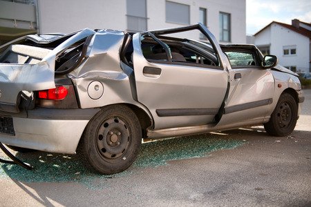 Completely wrecked silver hatchback sedan car with a flattened roof, shattered windows and buckled coachwork standing upright in the road surrounded by shattered glass shards following an accidentのeditorial素材