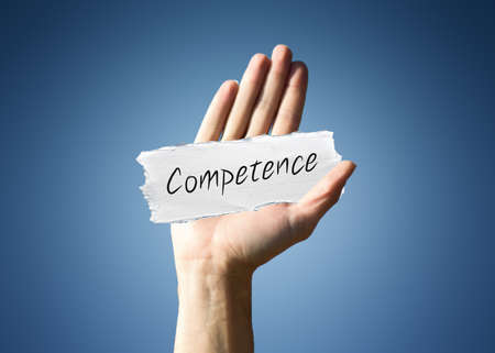 Man holding up a scrap of white paper with the word - Competence - in script, close up of his hand...