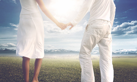 Rear view midsection of a couple in love wearing summer white clothes while holding hands on a green field with mountain range on the horizon, under the dazzling light of a bright skyの写真素材