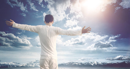 Man celebrating nature and his freedom standing with his back to the camera with wide outspread arms against a blue cloudy sky over snowy mountains with a bright sunburst and lens flareの写真素材