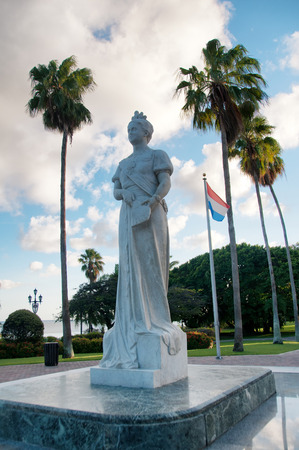Statue of Queen Wilhelmina in Oranjestad, Aruba with the Dutch flag standing in a square surrounded by tropical palm trees under a sunny cloudy blue summer skyの写真素材