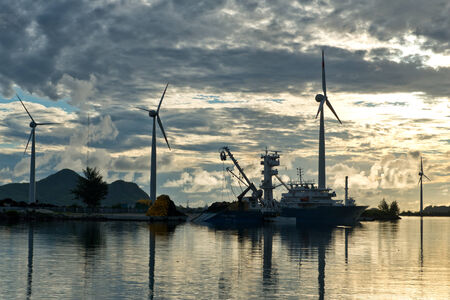 Row of wind turbines silhouetted at sunset at the edge of a tranquil sea with reflections with a ship sailing past on a cloudy evening in a sustainable energy conceptのeditorial素材