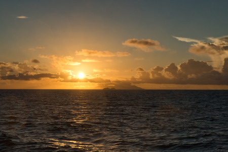 Fiery tropical ocean sunset over a calm blue sea as the glowing orange sun drops below the horizon and low cloud coverの写真素材