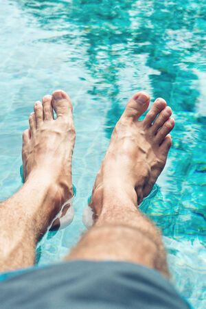 Close up bare male feet resting on crystal clear water at the relaxing swimming pool.