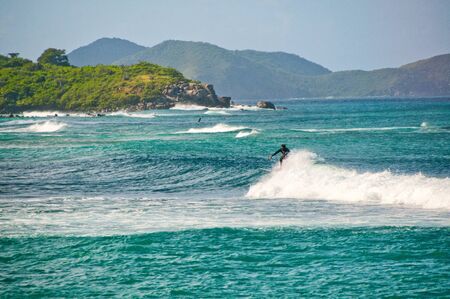 Surfer riding a wave at Tortola in the Leeward Islands in the Caribbean against a mountain backdrop with lush tropical vegetationの写真素材