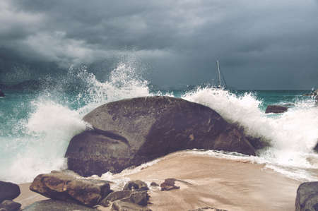 Wave breaking over a rock on a sandy tropical beach in a cloud of spray under a cloudy sky, close up view symbolic of summer vacationsの写真素材
