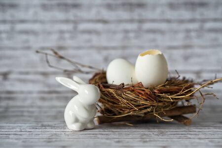 Close up Cute White Bunny Figurine with Whole and Broken Eggs in a Nest on Top of a White Wooden...