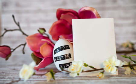 Easter Egg with Blank Vertical Card Surrounded by Colorful Spring Blossoms on Rustic Backgroundの写真素材