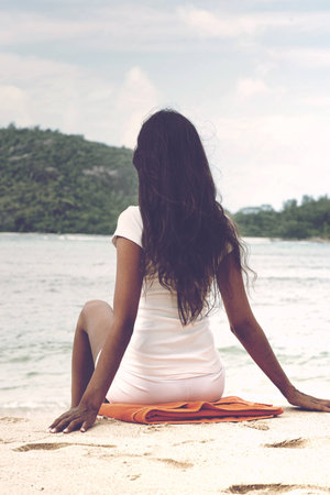 Close up Rear View Shot of Long Hair Woman in White Casual Outfit Sitting on White Beach Sand with Orange Towelの写真素材