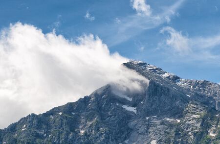 Snow-capped rocky mountain peak partially obscured by white cloud beneath blue sky