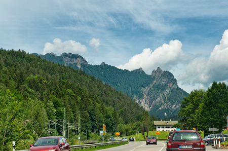 Wine coloured cars driving on road beside tree covered mountains beneath a cloudy blue skyのeditorial素材