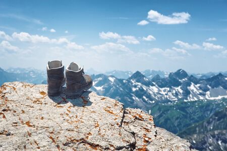 Pair of hiking boots on an alpine summit balanced on the edge of a rocky cliff overlooking distant alps with scattered snow on a sunny summer day, Hochvogel, Germanyのeditorial素材