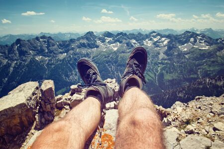 Climber in hiking boots on a rocky ledge overlooking the peaks of the Alps at Hochvogel, Germany, personal perspective looking down his legs to the viewの写真素材