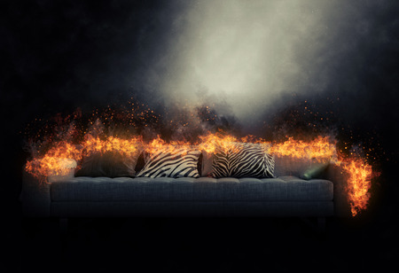 Large generic three-seater sofa engulfed in burning flames in a smoky dark atmosphere with a beam of light shining on it and copyspaceの写真素材