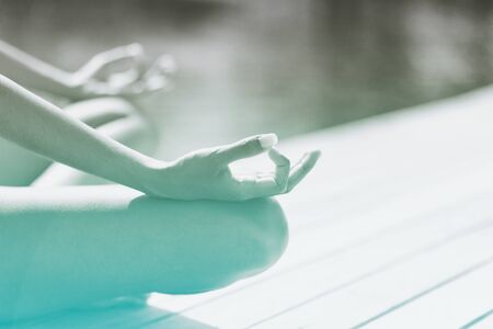 Young woman meditating on a wooden jetty or deck overlooking tranquil green-blue water, close up view of her hands and crossed legs in the lotus positionの写真素材