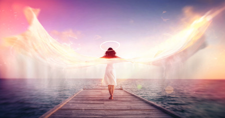Female angel standing barefoot on a jetty overlooking the ocean with wings in the form of billowing white fabric with motion blur with a halo and colorful sun flare effects, conceptual spiritual imageの写真素材