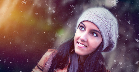 Attractive young Indian woman in a knitted winter hat watching the snow falling with a smile of pleasure and wonderment over a blurred outdoor background with copy spaceの写真素材