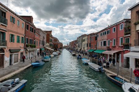 Canal scene on Murano, Venice, Italy with the sidewalks lined with shops and tourists and boats moored along the length of the canal on a cloudy blue sky dayの写真素材
