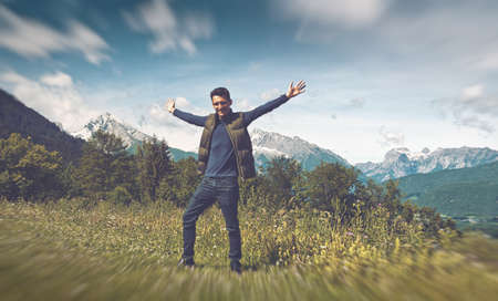 Full length of 20s man wearing dark jeans, navy top and gillet with arms out-stretched standing on grass. Snow-capped mountain vista and cloudy blue sky backdropの写真素材