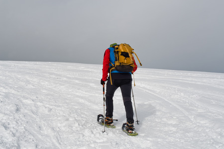 Rear view of unidentifiable cross country snowshoe trekker traveling upward on mountain into dark gray storm cloudsの写真素材