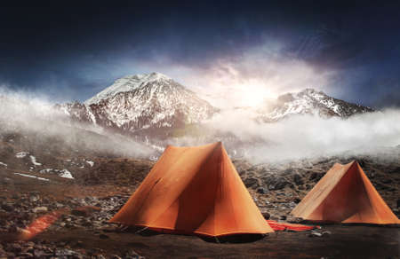 Two tents pitched below snow capped mountains at sunrise with a bright sunburst over the tops of the peaks and early morning mist clinging to the landscapeの写真素材