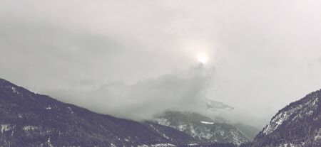 Panoramic view of light from sun barely peaking from clouds above beautiful tall mountain rangeの写真素材