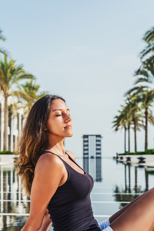 Serene young woman meditating in the summer sun in front of a palm tree lined pool at a tropical...