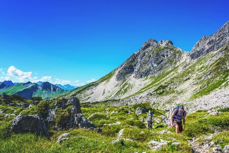 Hikers traveling a path surrounded by green grass and stones under blue sky in the German Alps
