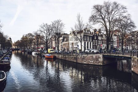 Water canal in Amsterdam with boats, bridge and historical canal houses surrounded by bare trees, with cloudy sky reflected in waterのeditorial素材