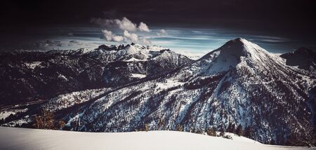 Dark moody artistic alpine panorama with a tall snow-capped forested mountain peak and fresh white winter snow in the foreground surrounded by a heavy vignetteの写真素材